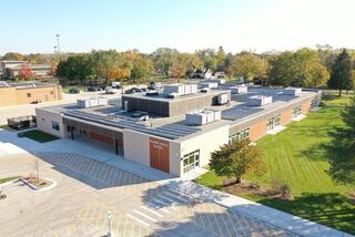 A modern building with a flat roof surrounded by green grass and trees, featuring a spacious parking area and multiple entrances.
