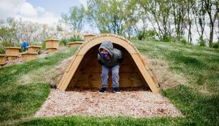 A child peeks out from a wooden play structure on a grassy hill, surrounded by nature and playful wooden installations.
