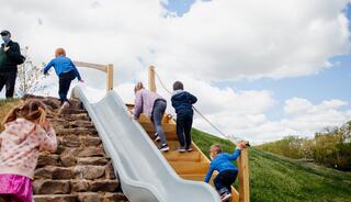 Children are climbing a stone staircase to a slide, enjoying a sunny day with fluffy clouds in the background.