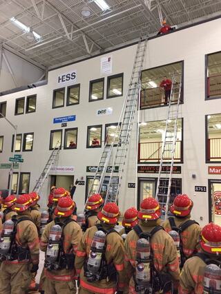 Firefighters in gear prepare for a drill involving ladders and a multi-level training facility, focusing on emergency response techniques.