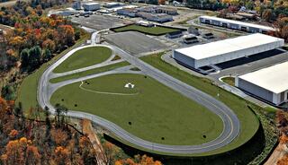 An aerial view of a racing track surrounded by warehouses and autumn foliage, featuring winding paths and expansive green spaces.