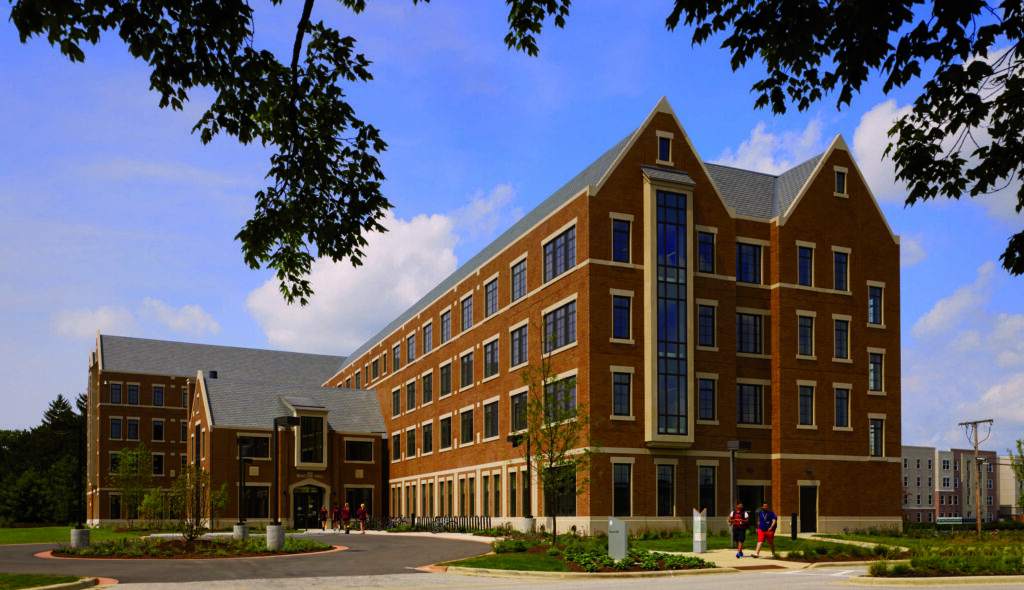 A modern, multi-story brick building with large windows, surrounded by greenery and pathways, indicative of a campus setting with people walking.