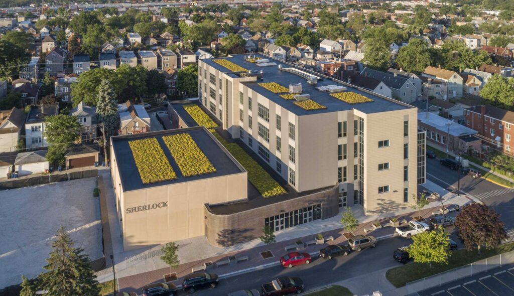 A modern multi-story building features green rooftop gardens and is surrounded by residential areas and streets, showcasing urban development.