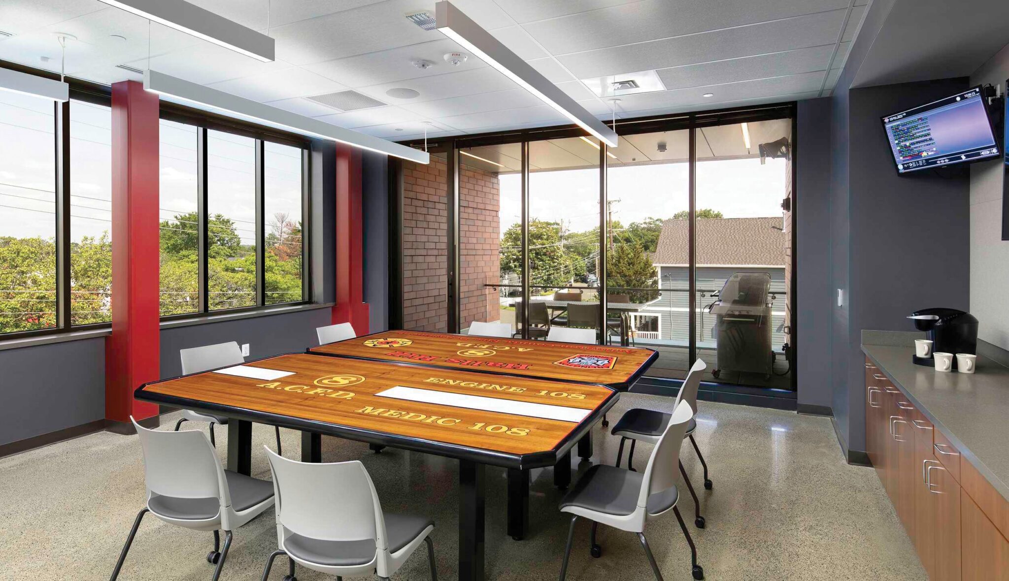 A dining room featuring a large table with fire station logo, surrounded by chairs and large windows offering a view of greenery outside.