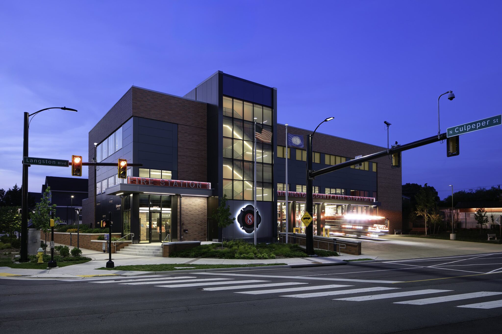 A modern building with a mix of brick and glass, illuminated at dusk, stands at an intersection with traffic lights and signage.
