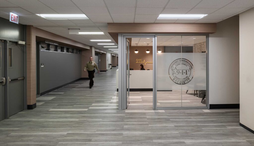 A modern office space with glass walls, warm lighting, and a spacious hallway featuring a person walking in the background.