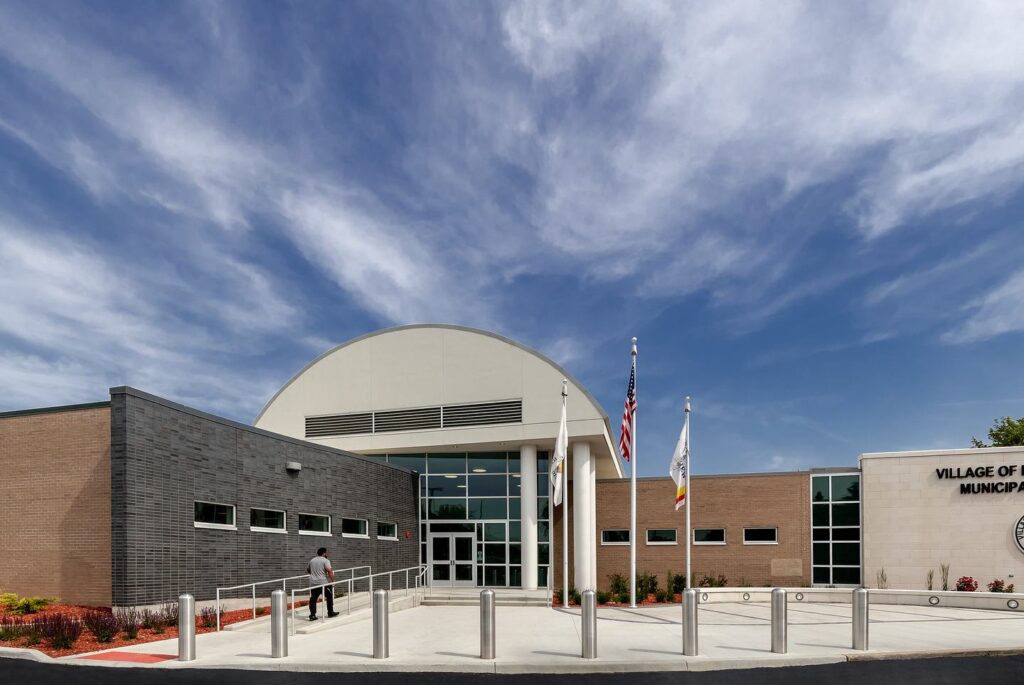 A modern municipal building features a curved roof, flagpoles, and landscaped surroundings under a bright, cloudy sky.