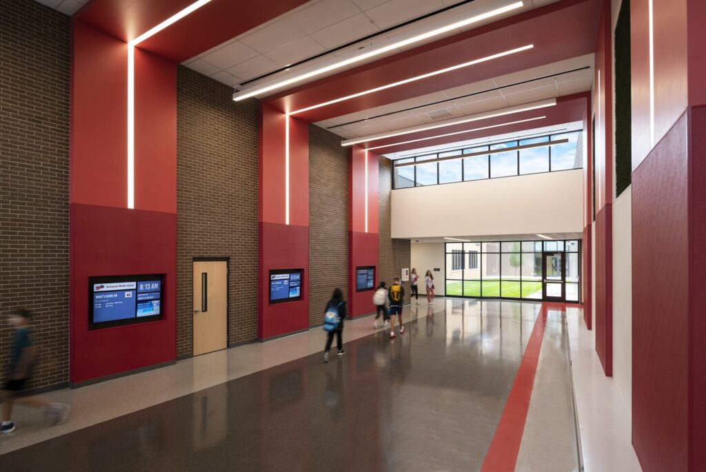 A spacious hallway features red accents, large windows, digital displays, and students walking, creating a vibrant, modern educational environment.