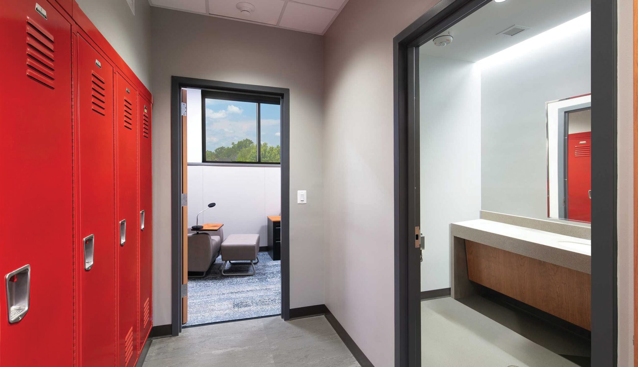 A bunk room features red lockers on one side, with a doorway leading to a room containing a bed and a bright, open bathroom.