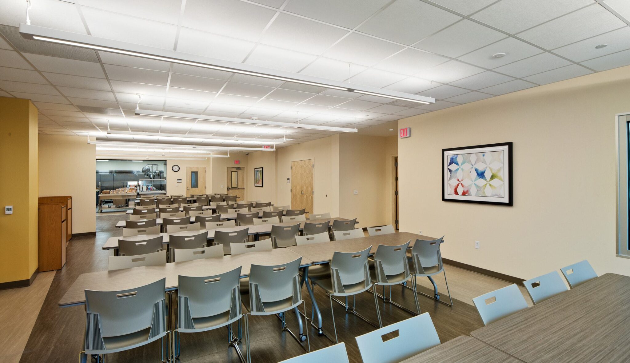 A spacious room with rows of tables and chairs, brightly lit, featuring a kitchenette and a decorative framed artwork on the wall.
