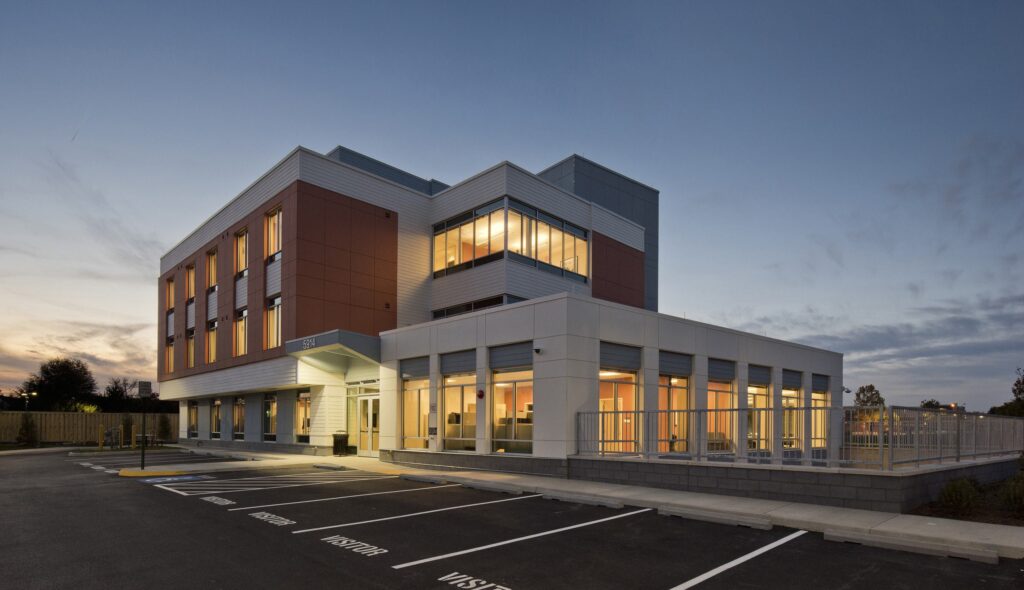 A modern building features large windows and a mix of brick and sleek design elements, surrounded by a parking lot at dusk.