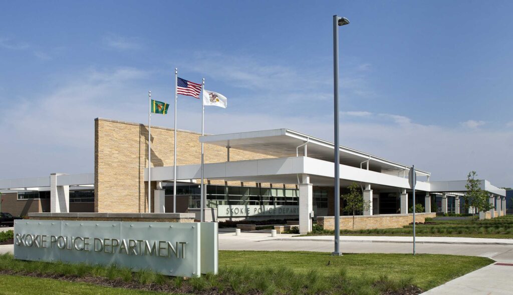 A modern building with a sleek design showcases flags and a prominent sign, set against a clear blue sky and green landscaping.