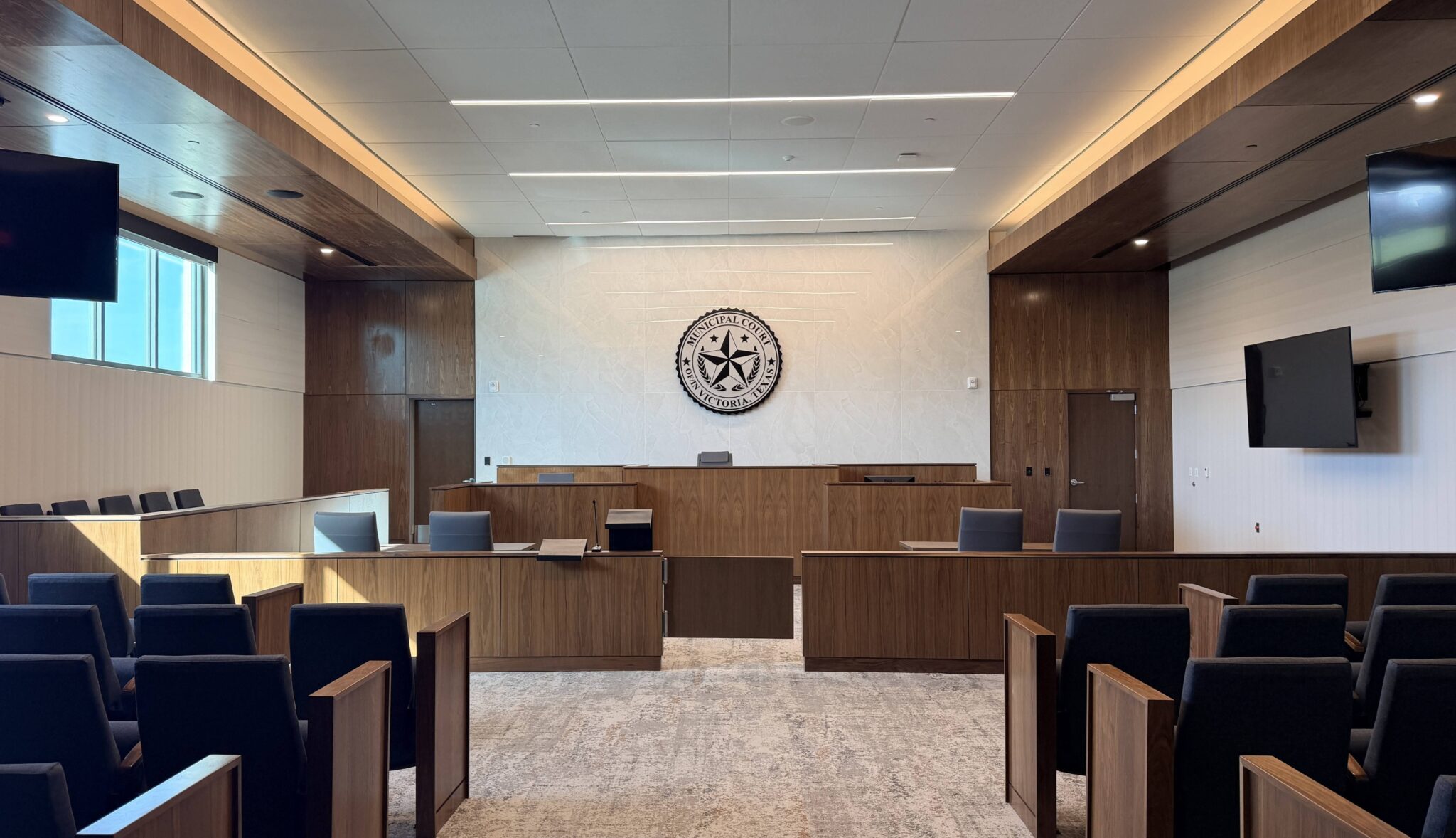 A modern courtroom features wooden furnishings, multiple screens, and a decorative emblem on the wall, emphasizing a professional atmosphere.