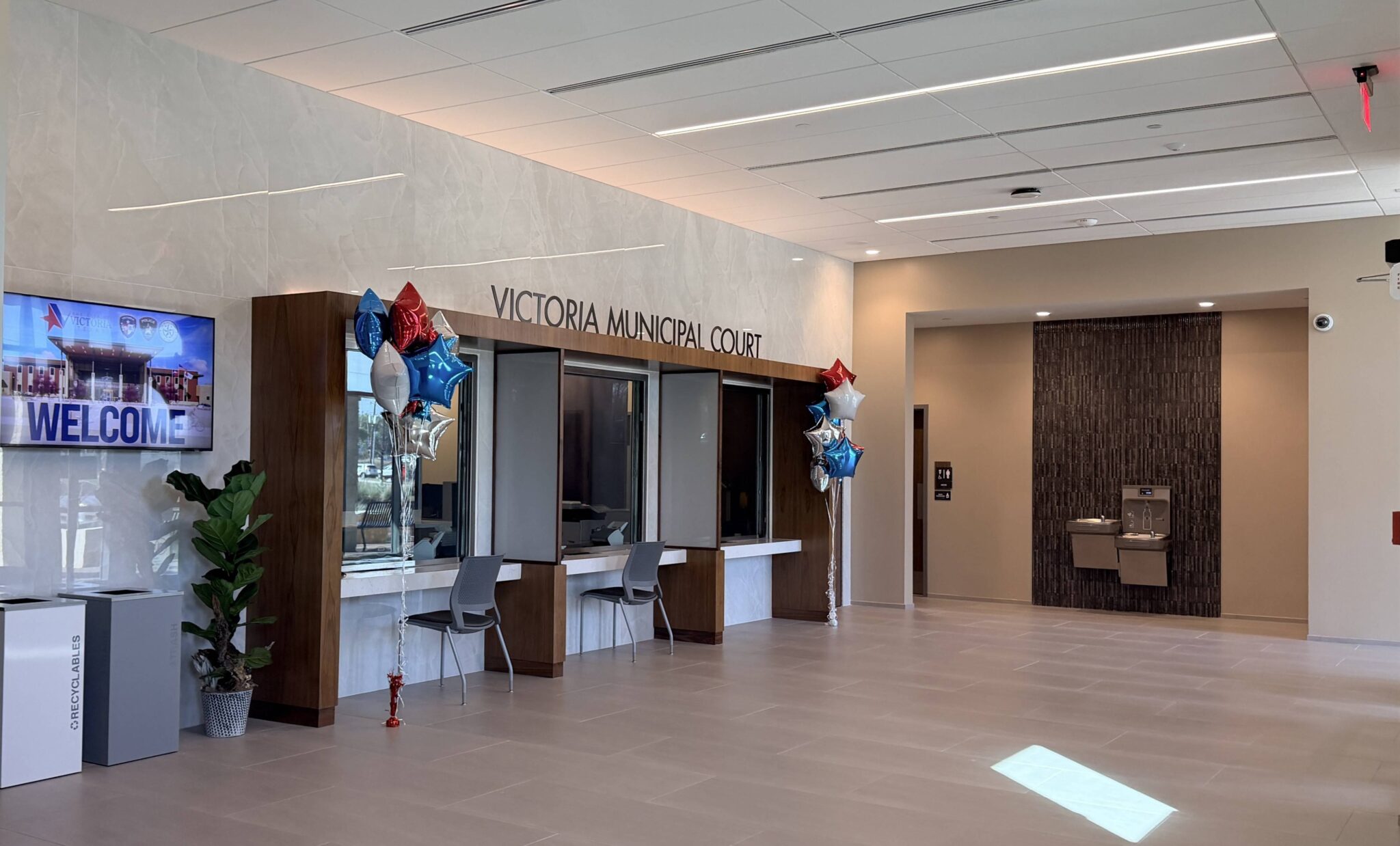 A modern municipal court lobby features a welcoming sign, service windows, seating, decorative balloons, and a water fountain.