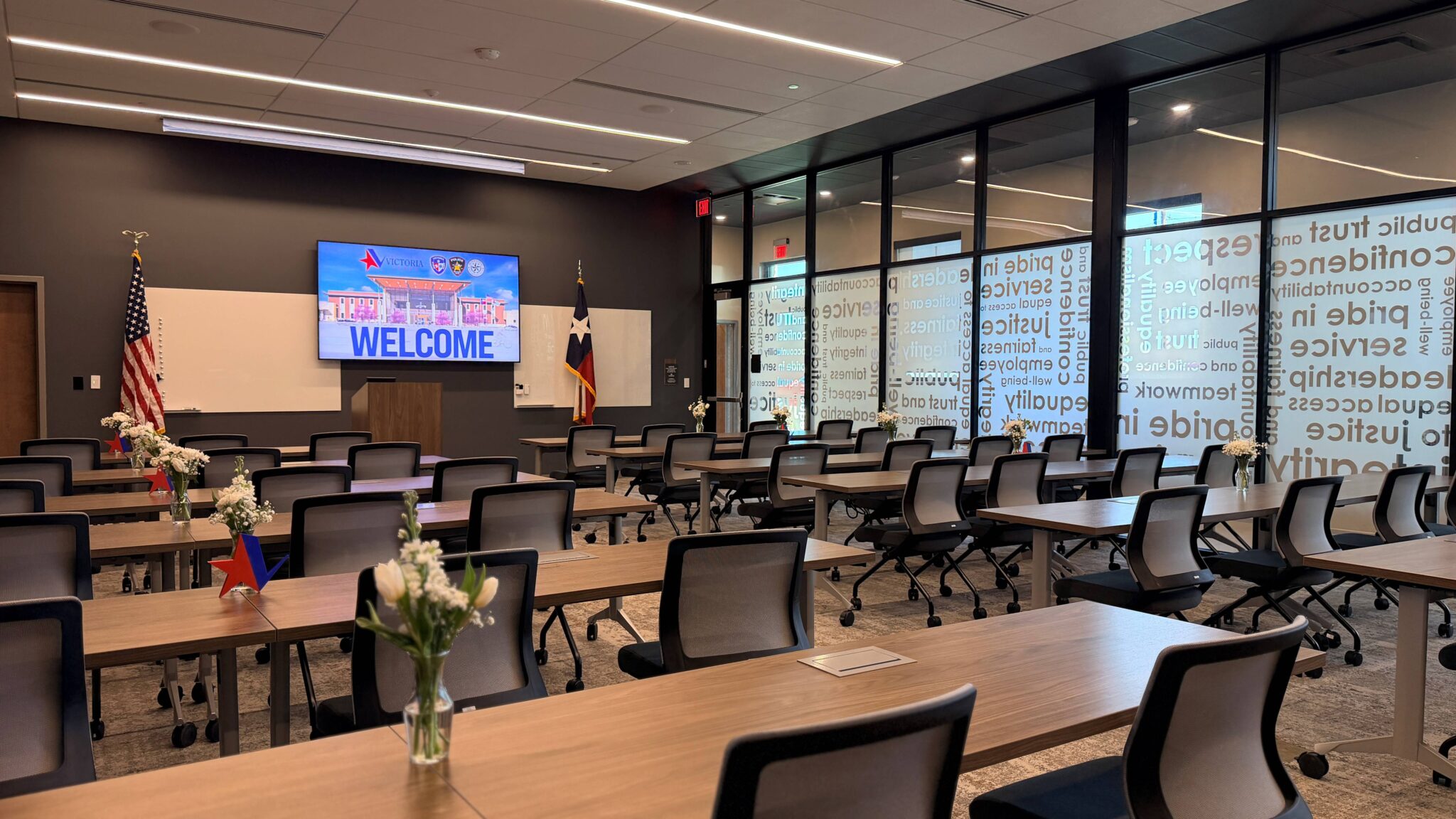 A modern classroom setup features rows of tables and chairs, with a large screen displaying "WELCOME," flags, and decorative flowers.