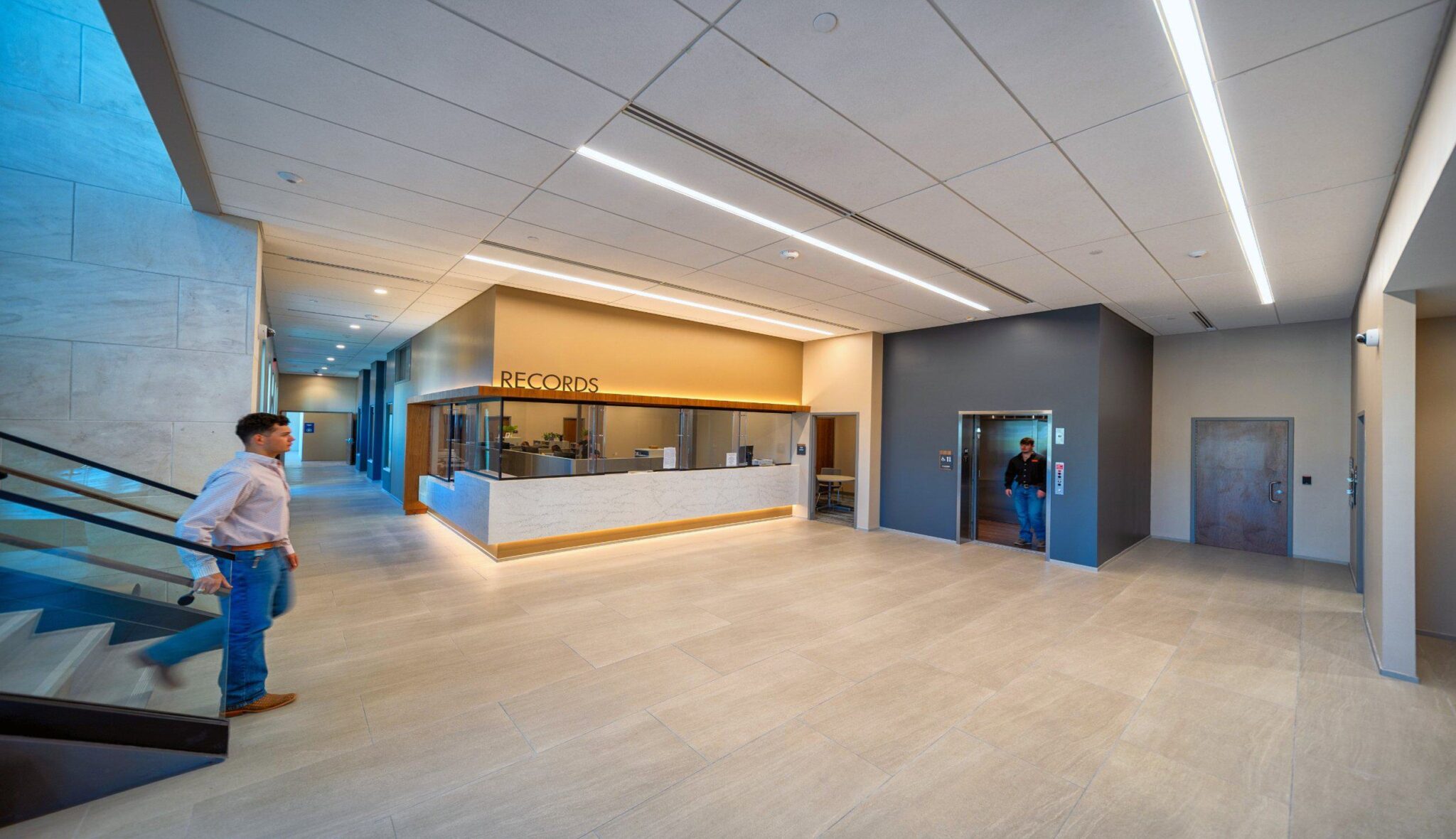 A modern, spacious reception area features a sleek records desk, elevators, and ample natural light, with neutral tones and stylish finishes.