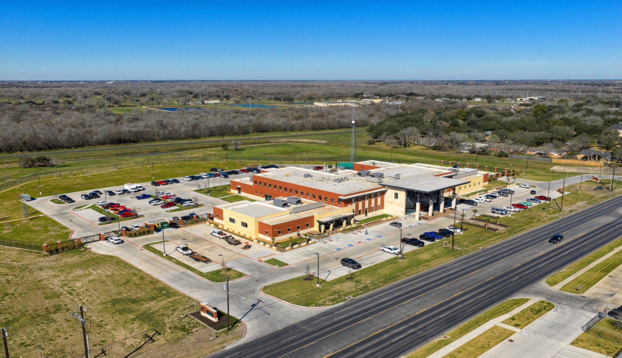 Aerial view of a modern building with multiple vehicle parking spaces, surrounded by lush green fields and trees under a clear blue sky.