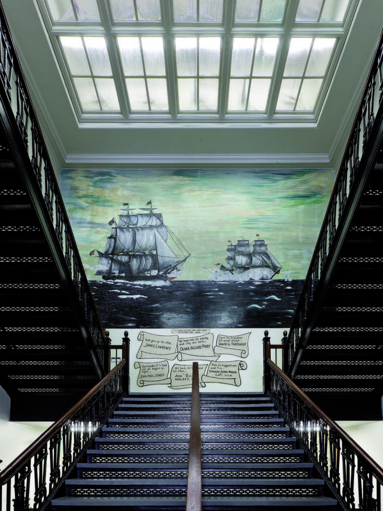 A grand staircase leads up to a vibrant mural of sailing ships, illuminated by skylights, with historical texts displayed below.