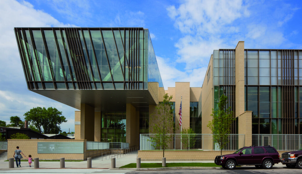 A modern architectural structure features a striking glass canopy, bold lines, and greenery, reflecting contemporary design elements.