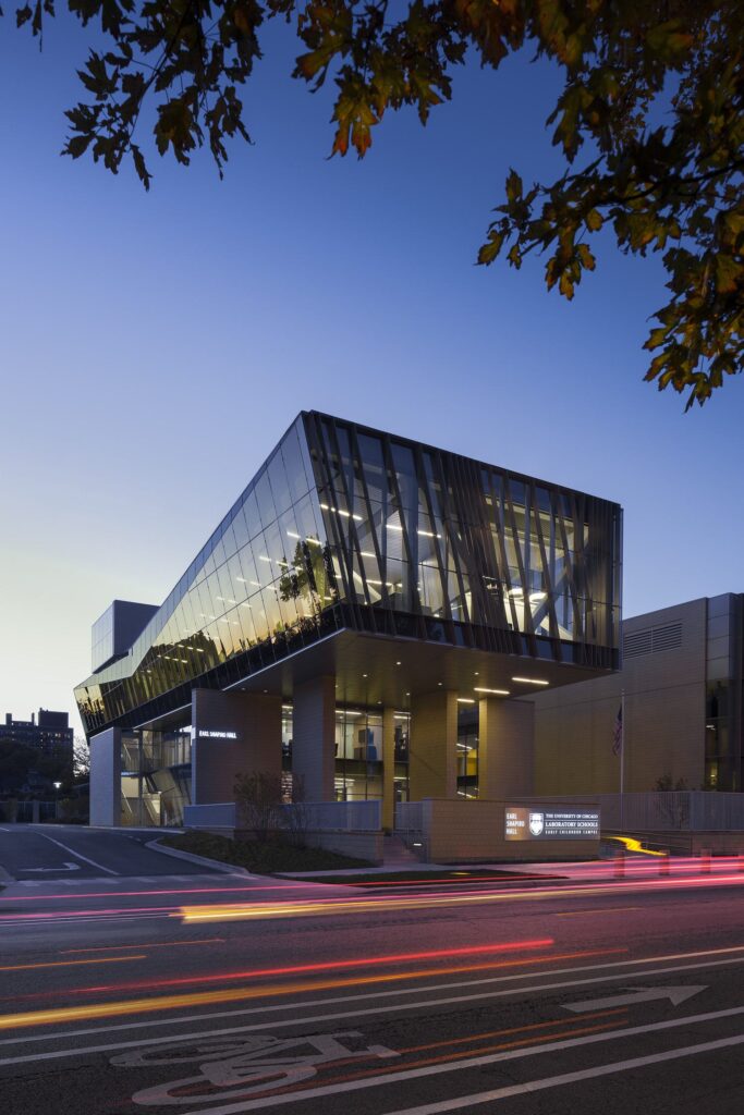 A modern architectural building featuring glass panels and angular design, illuminated against a twilight sky with light trails from passing vehicles.