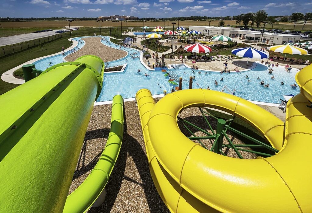 Brightly colored water slides curve over a pool area filled with sun umbrellas and families enjoying a sunny day at a water park.