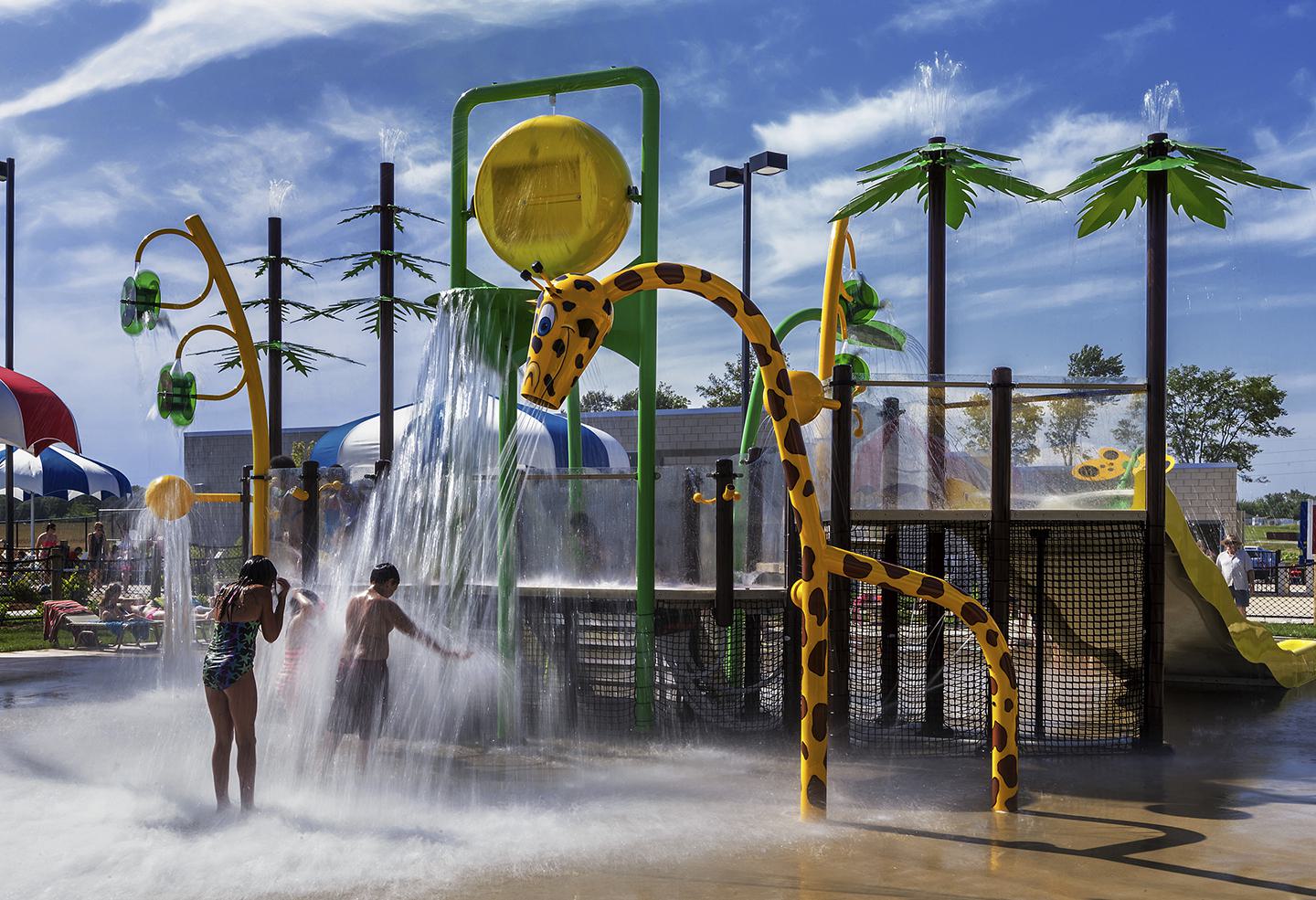 A colorful splash pad features a giraffe structure, palm trees, and kids enjoying water play under a sunny sky. Fun summer vibes!