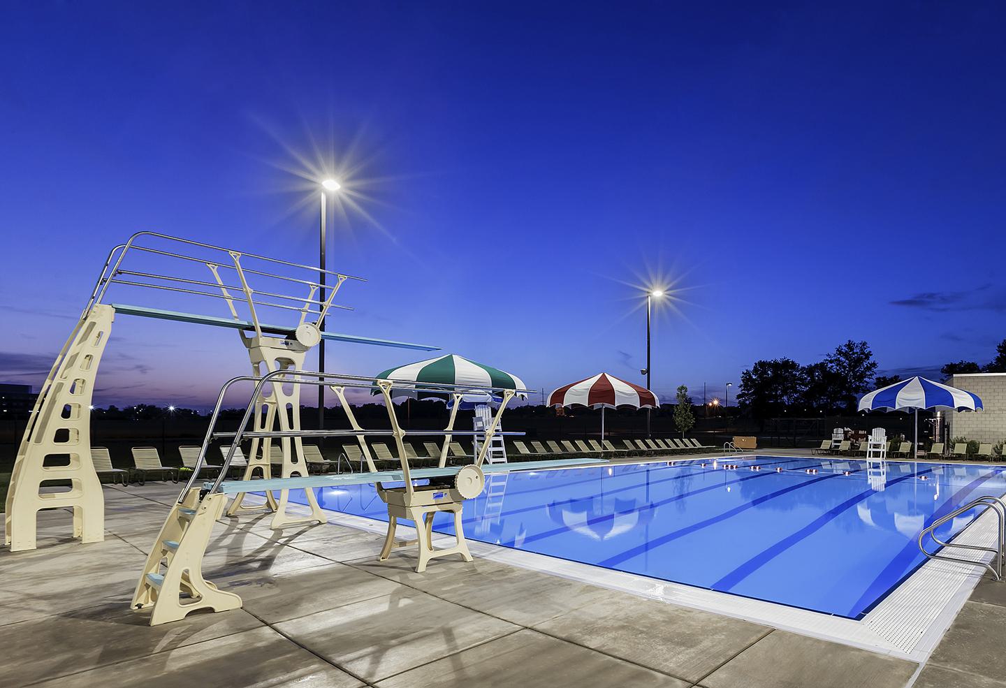 A tranquil pool area at dusk, featuring a diving board, striped umbrellas, and bright lights illuminating the calm water.