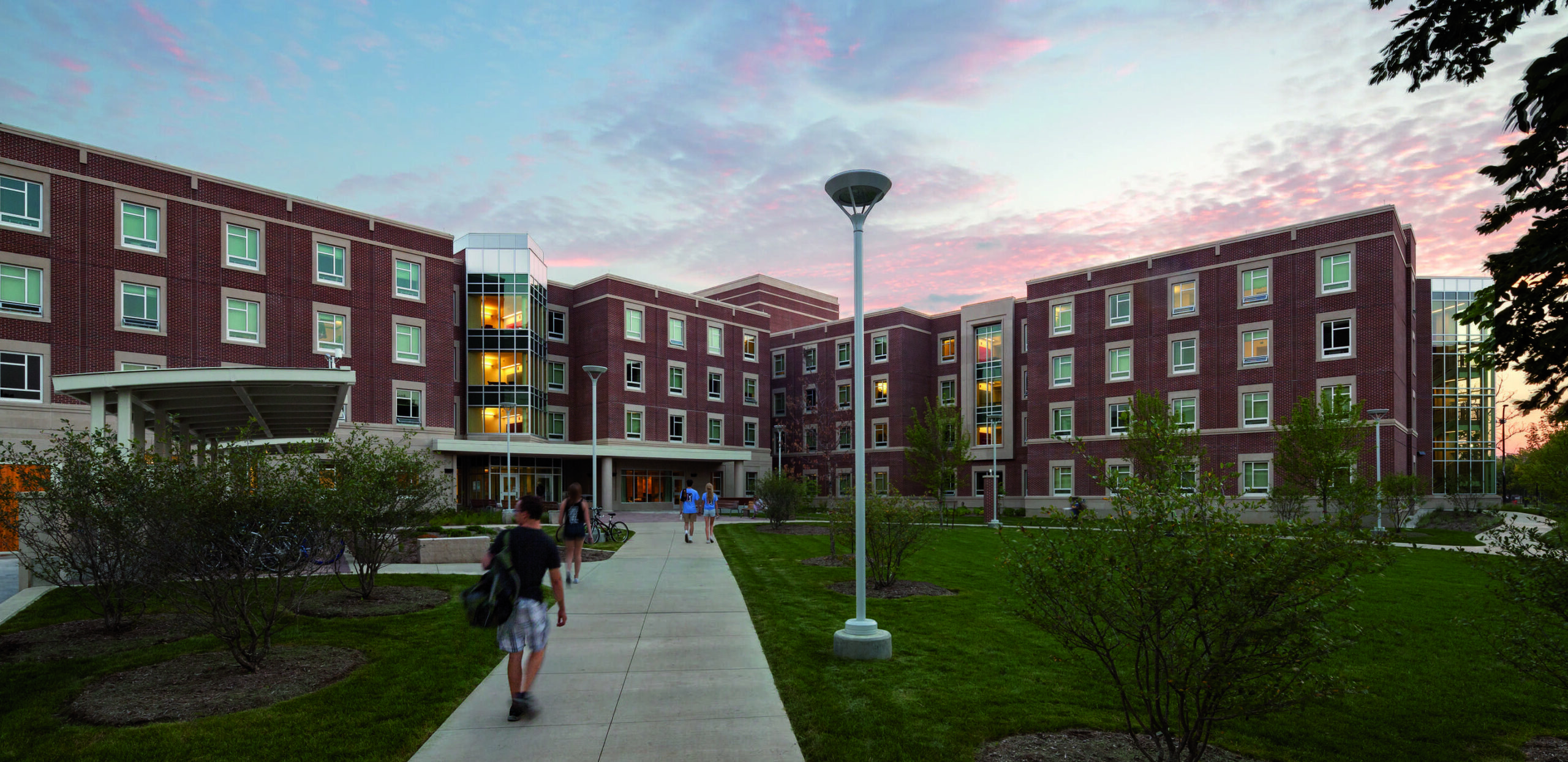 A modern brick building features a walkway, greenery, and students walking, all under a vibrant, colorful sky at dusk.
