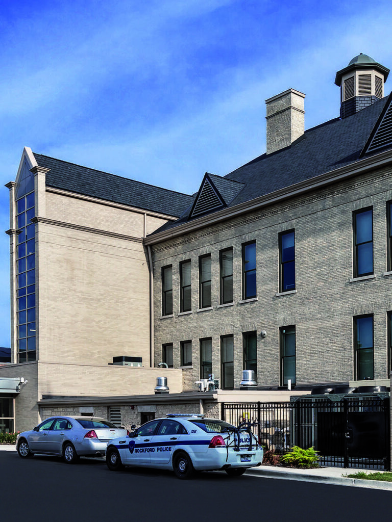 A modern building features brick architecture with a flat roof. Two police vehicles are parked nearby, set against a clear blue sky.