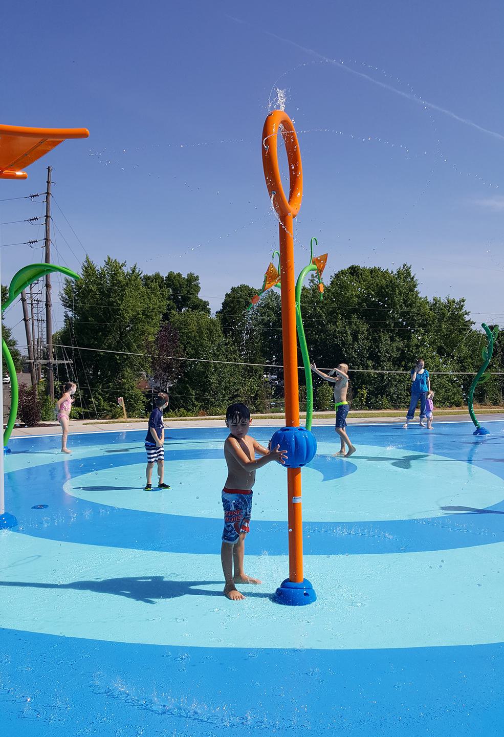 Children play in a colorful splash pad, some interacting with water features under a clear blue sky, surrounded by greenery.