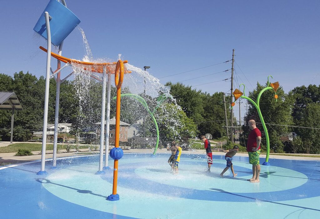 Children play in a colorful splash pad, enjoying water features under a clear blue sky while a guardian watches nearby.