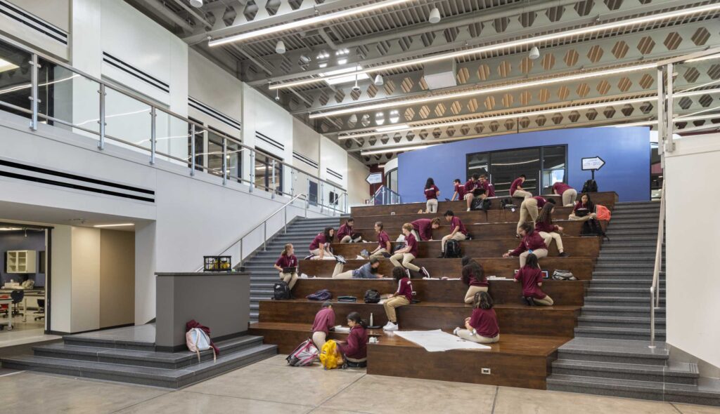 Students sit on large wooden steps in a modern learning space, engaged in various activities, with bright walls and ample natural light.