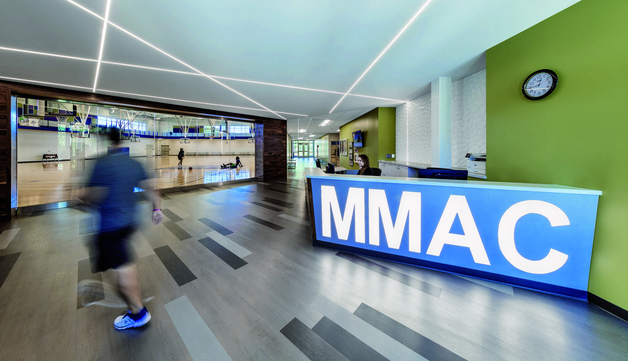 A bright entry lobby at Madison Meadows Athletic Center features a large "MMAC" sign at the reception desk, sleek flooring, and a glimpse of an active gym area in the background.
