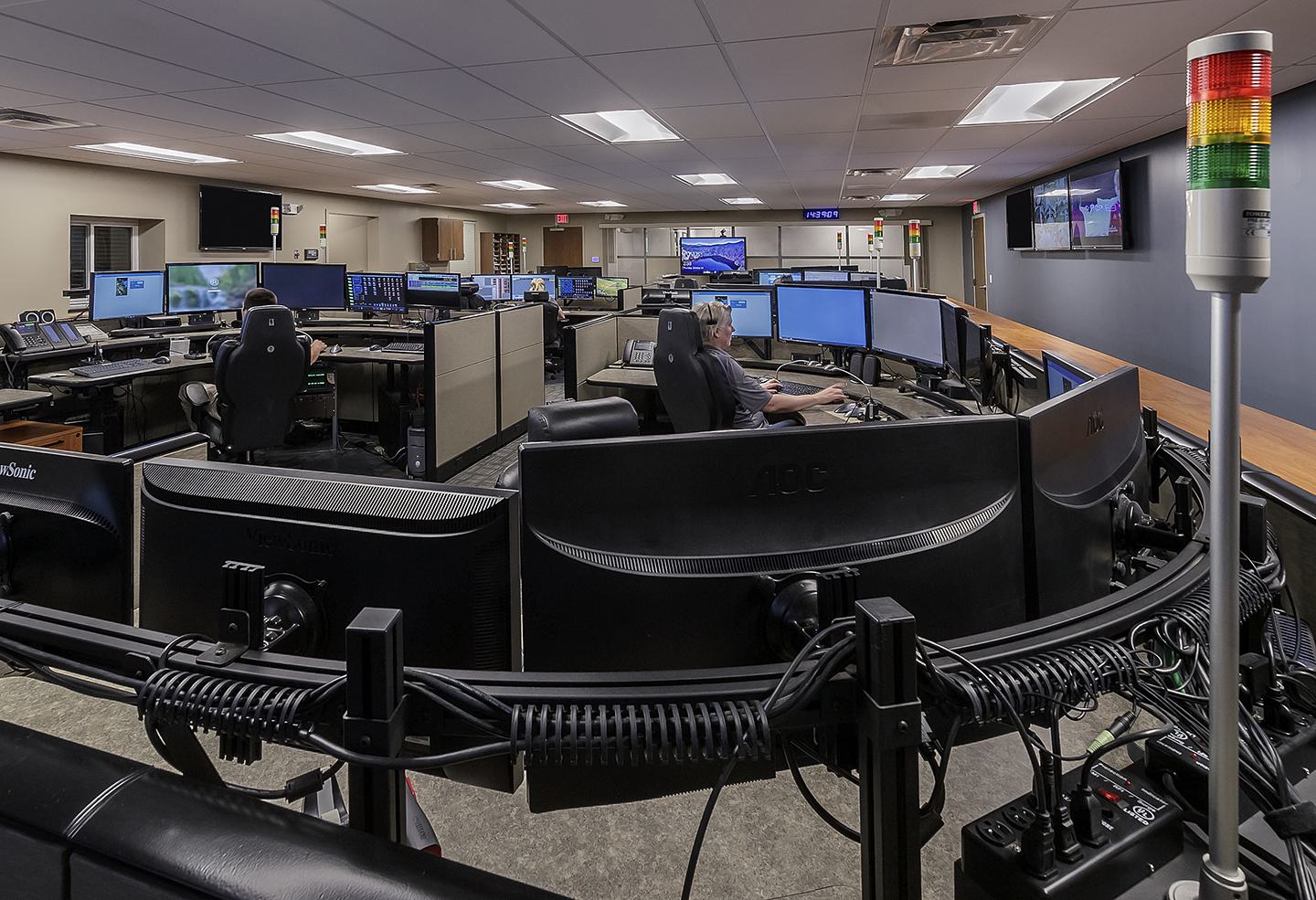 A modern control room features multiple screens, with an operator sitting at a central workstation, surrounded by monitoring equipment and staff.