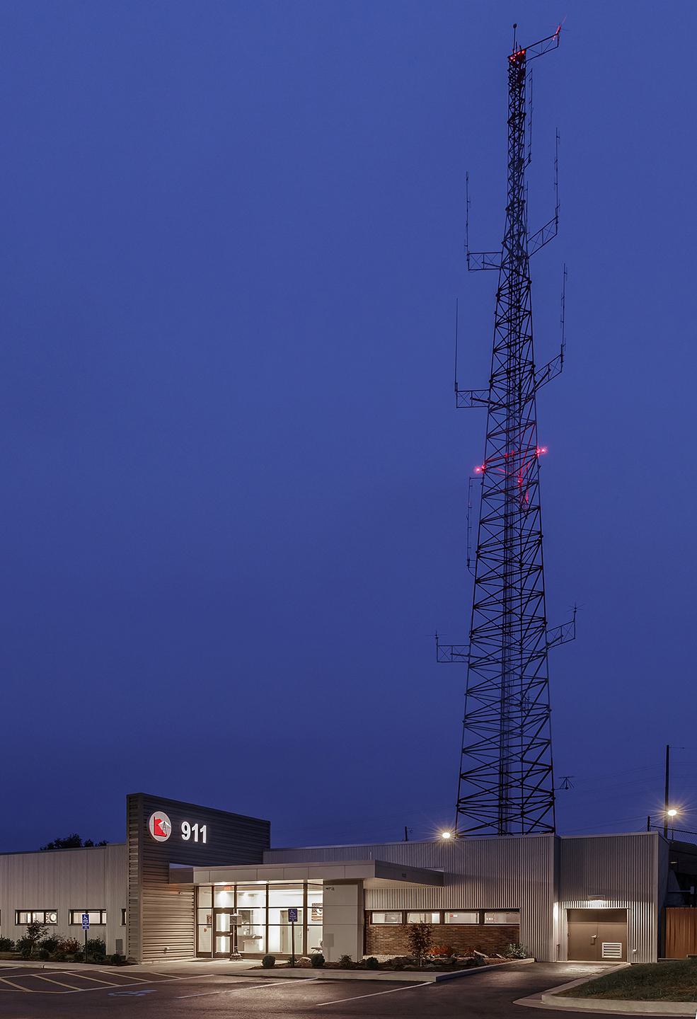 A modern emergency services building with a tall communication tower, illuminated under a blue twilight sky. The "911" sign is prominent.
