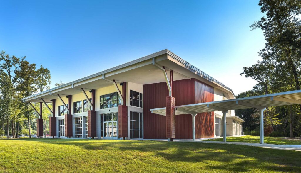 A modern building with large glass windows and a striking roof design, surrounded by lush greenery and tall trees.