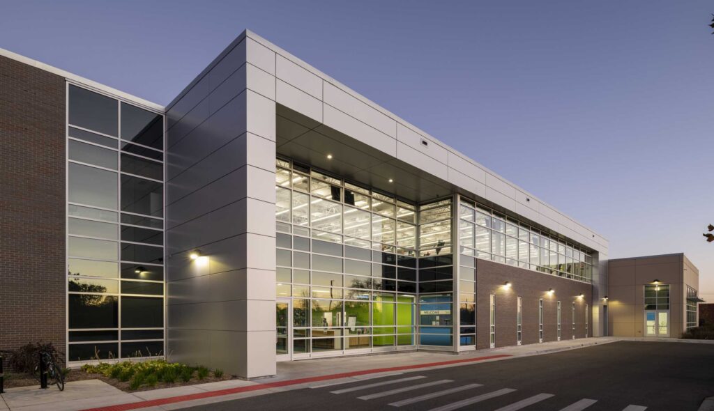 A modern building features sleek glass and metal architecture, illuminated at dusk, with a welcoming entrance and landscaped surroundings.