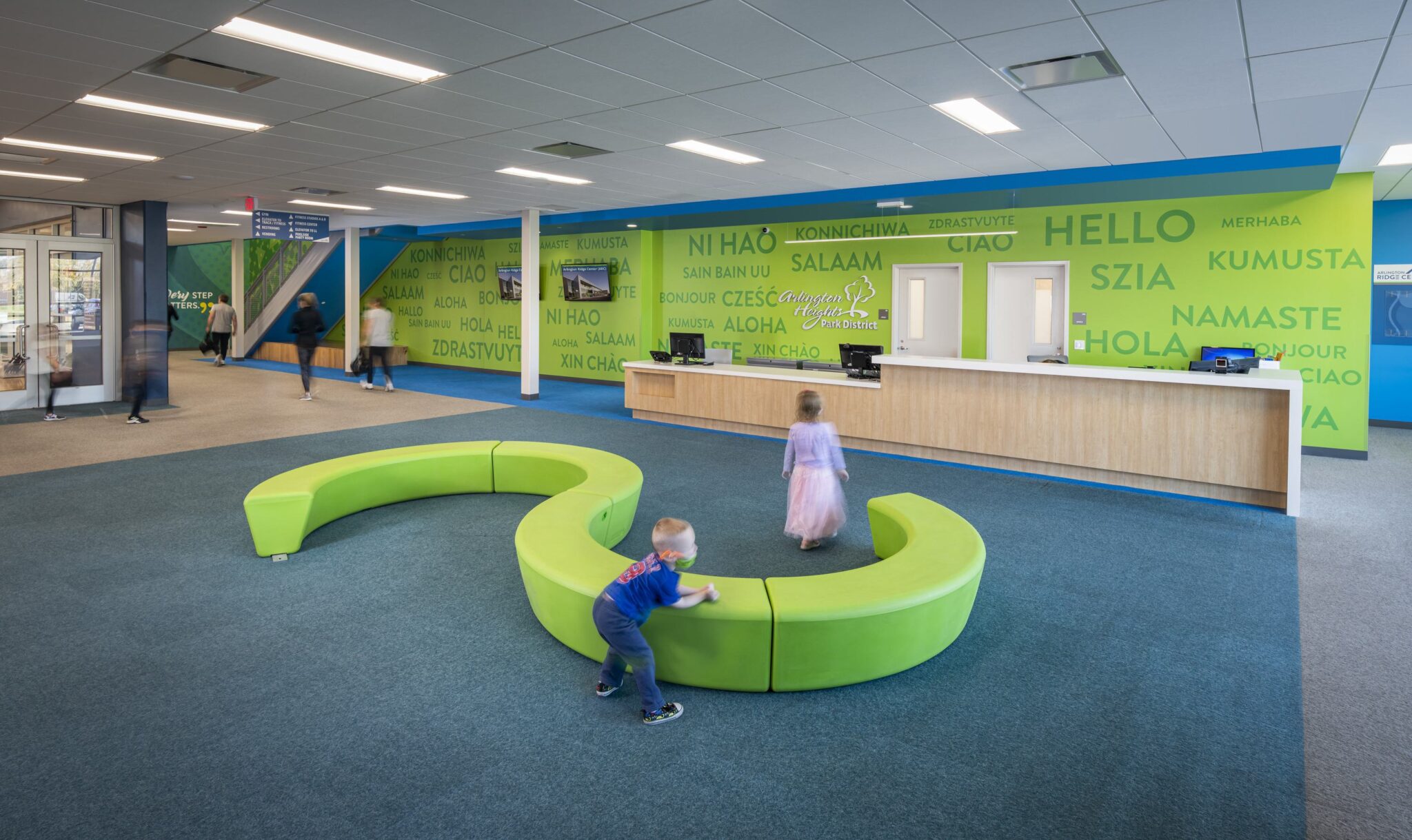 A modern space features a vibrant green seating area, with children playing and colorful greetings in multiple languages on the walls.