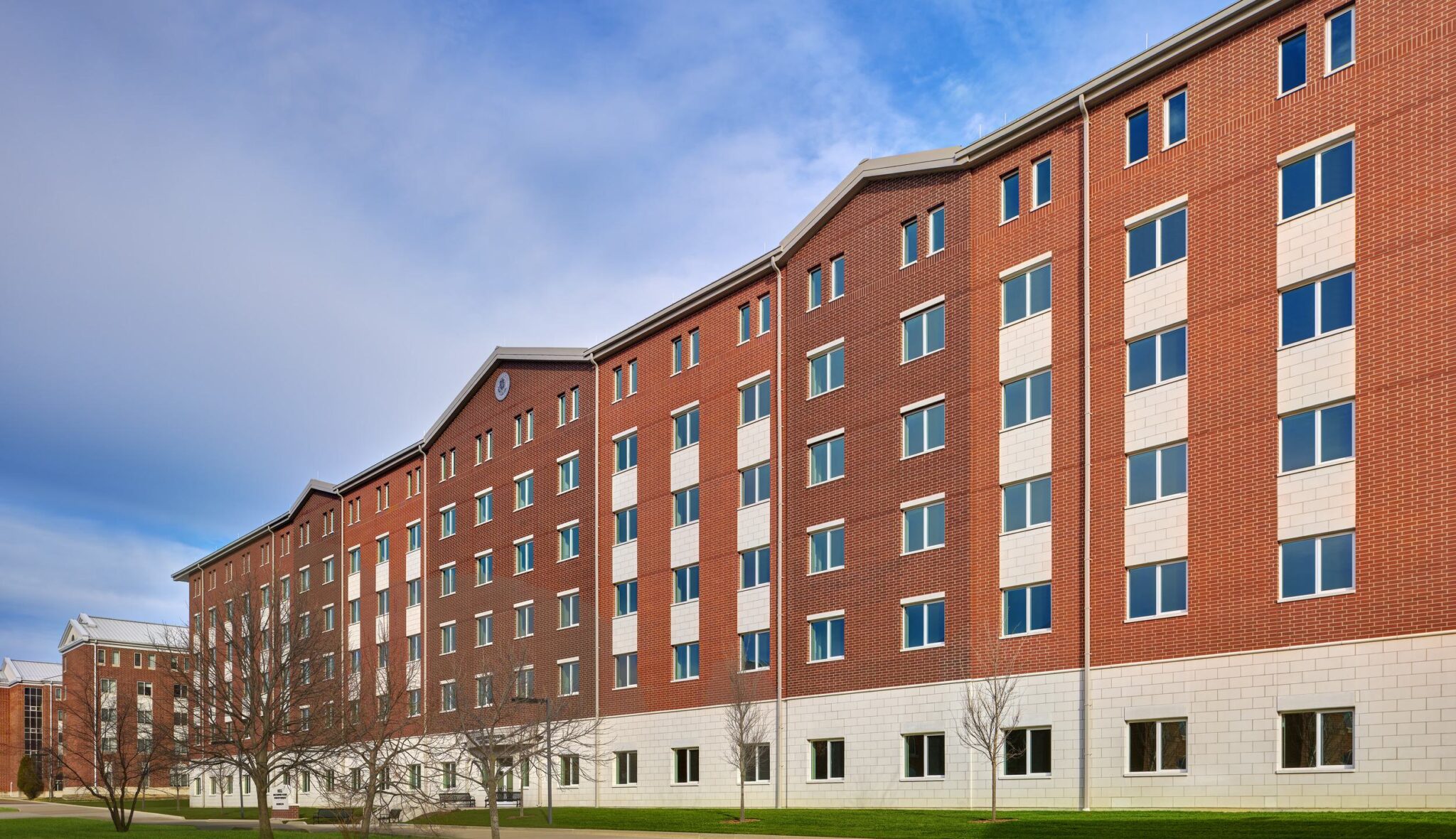 A large, modern brick building with multiple floors and windows, set against a blue sky, surrounded by a grassy area and trees.