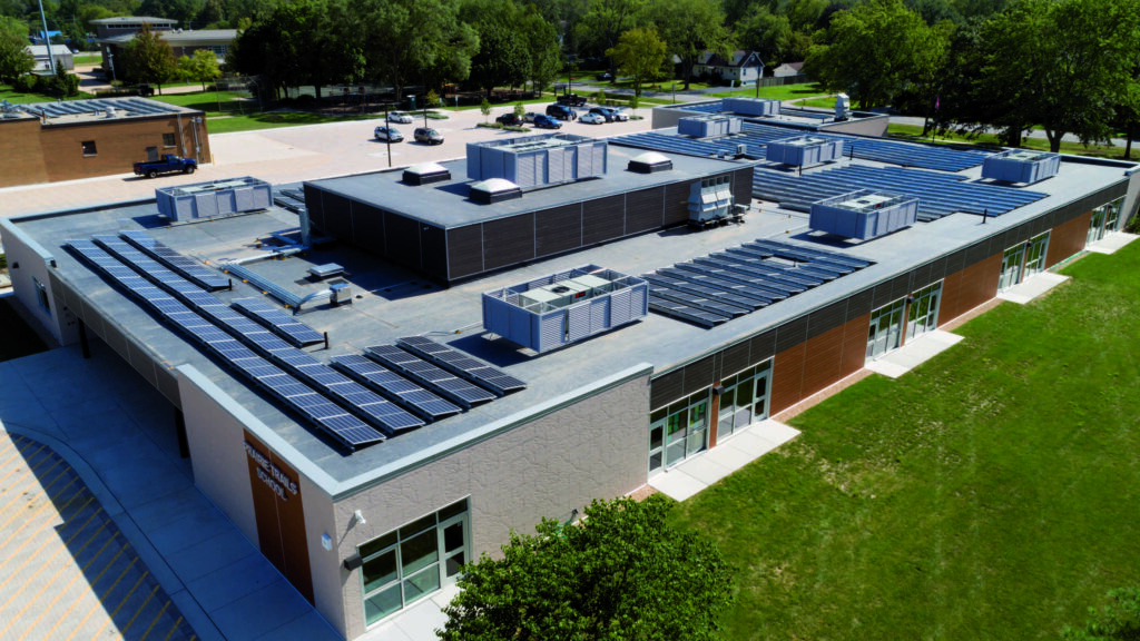 Aerial view of a modern building featuring multiple rooftop units and solar panels, surrounded by green landscape and parking area.