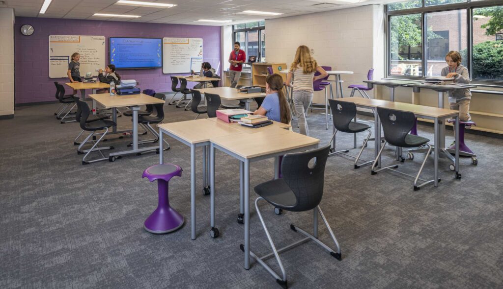 A modern classroom features flexible seating with students engaged in various activities, bright natural light, and interactive learning spaces.