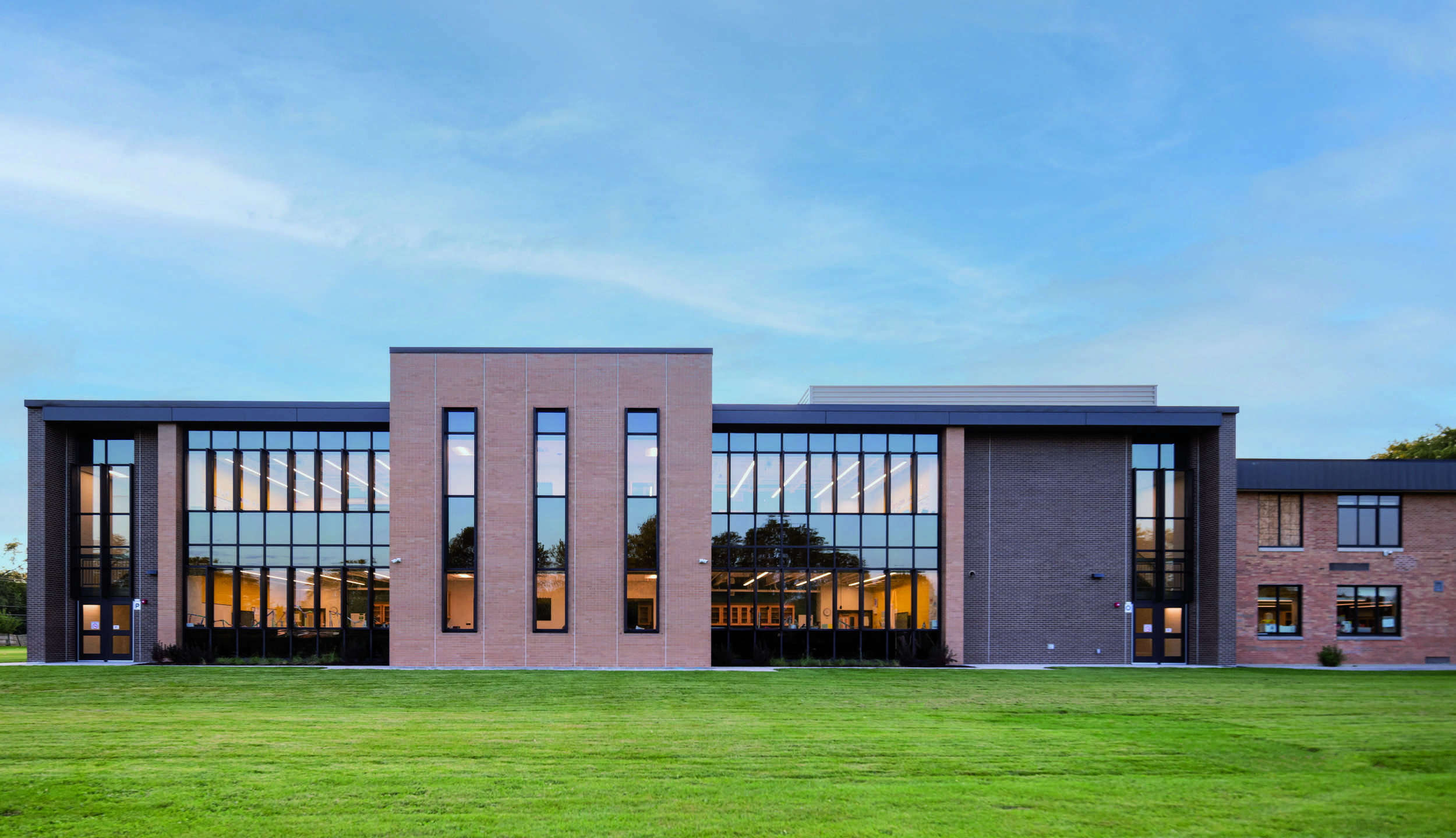 A modern, two-story building features large glass windows and a symmetrical design, surrounded by a well-kept grassy area under a clear sky.