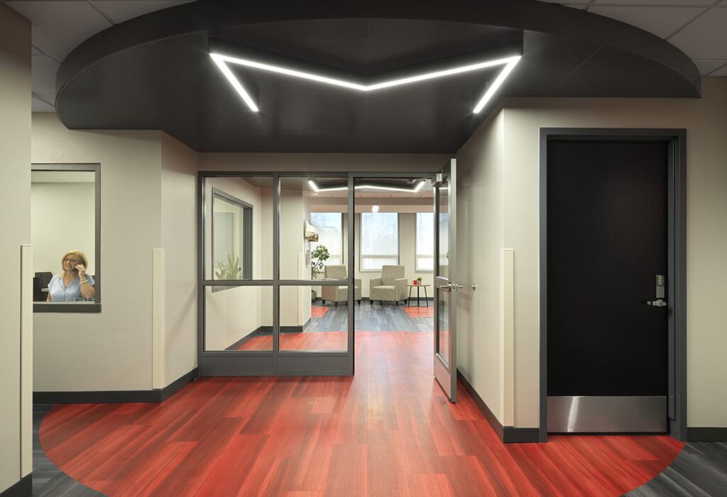 A modern hallway features red flooring, sleek lighting, glass partitions, and an inviting lounge area visible through large windows.