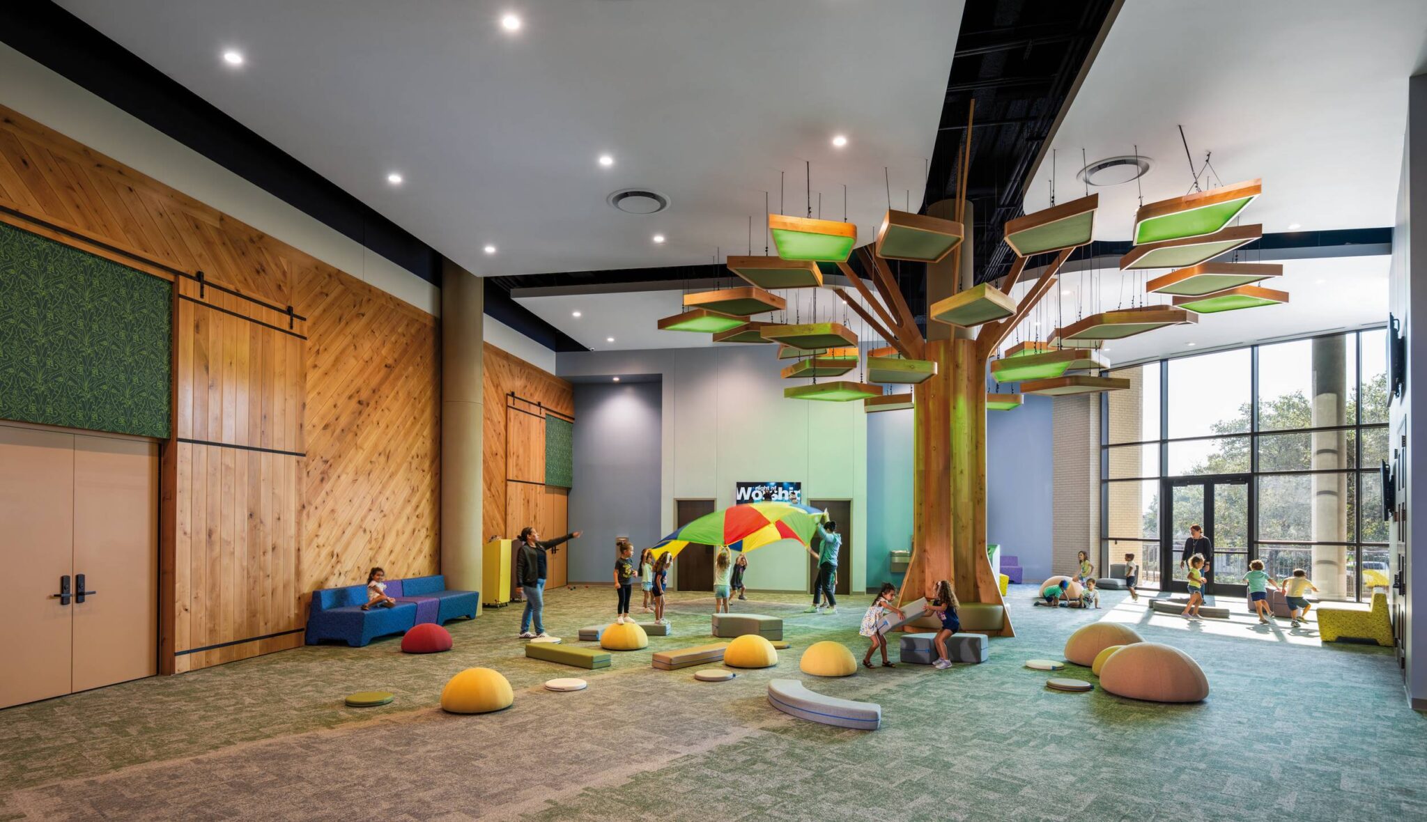 A spacious, playful room features a tree-like structure with colorful leaves, soft seating, and children engaging in various activities.