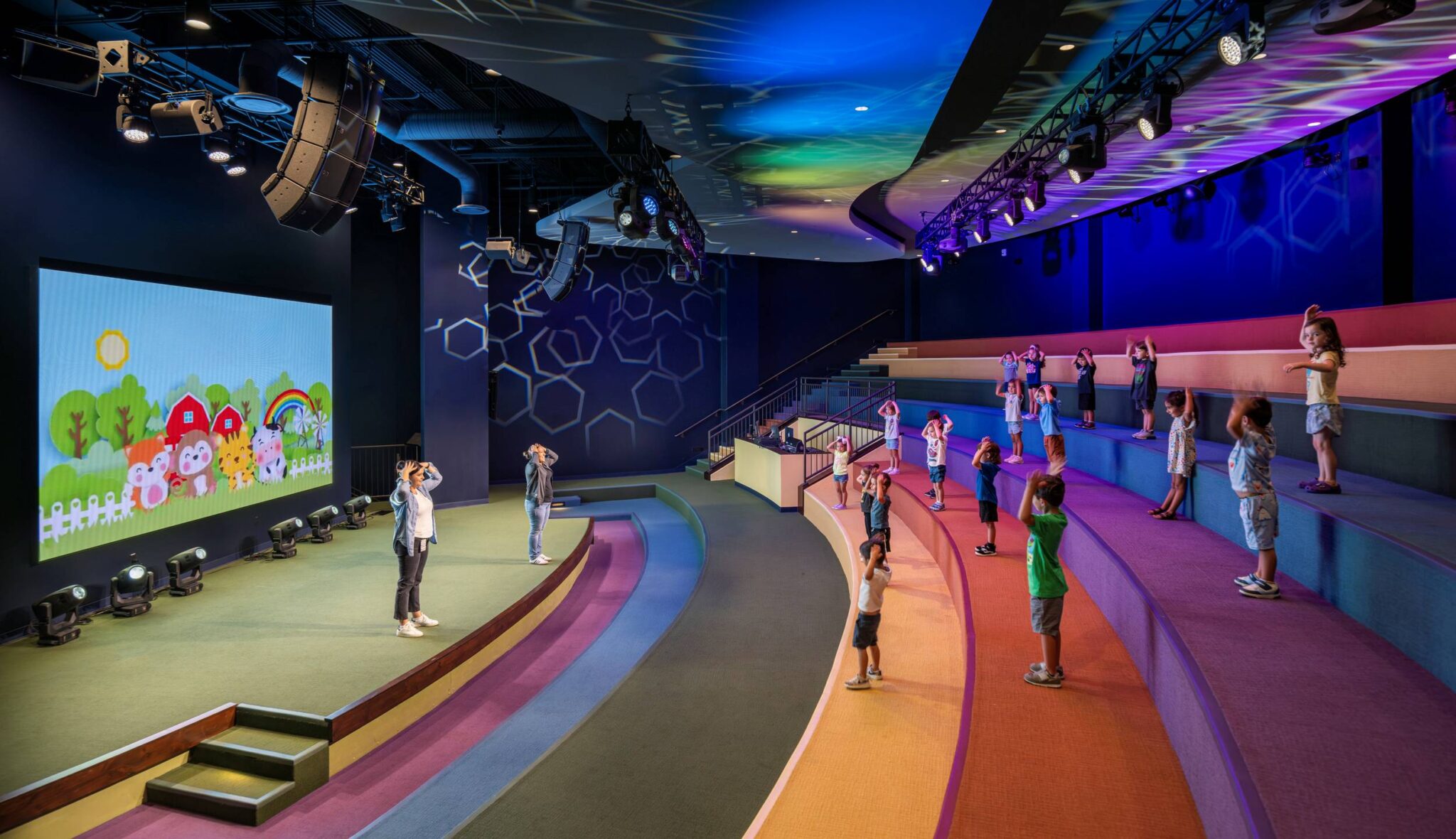 Children gather in a colorful, modern space, following a leader on stage, with vibrant visuals enhancing the lively atmosphere.