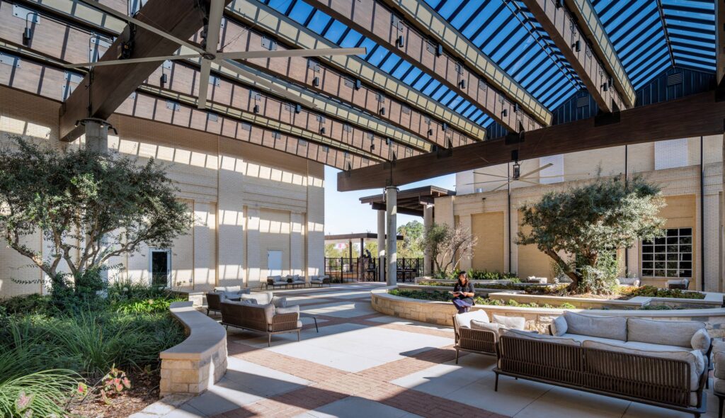 A modern, open courtyard features comfortable seating, lush greenery, and a striking glass roof, creating a serene outdoor space.