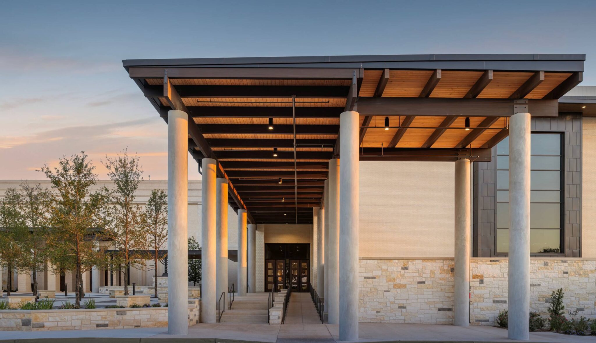 A modern building entrance features a wooden roof with steel columns, surrounded by landscaped greenery and a serene sky at dusk.