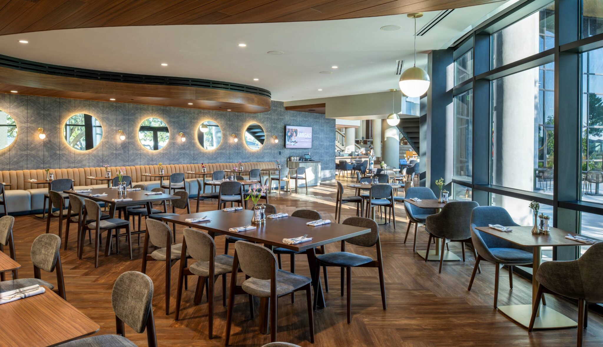A modern restaurant interior featuring wooden accents, stylish furniture, circular mirrors, and large windows creating a bright, inviting atmosphere.