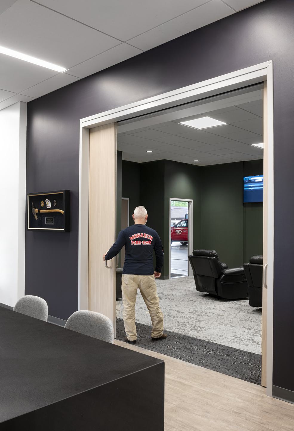 A person in a shirt walks through a doorway into a modern lounge area with recliners and a dark green wall.