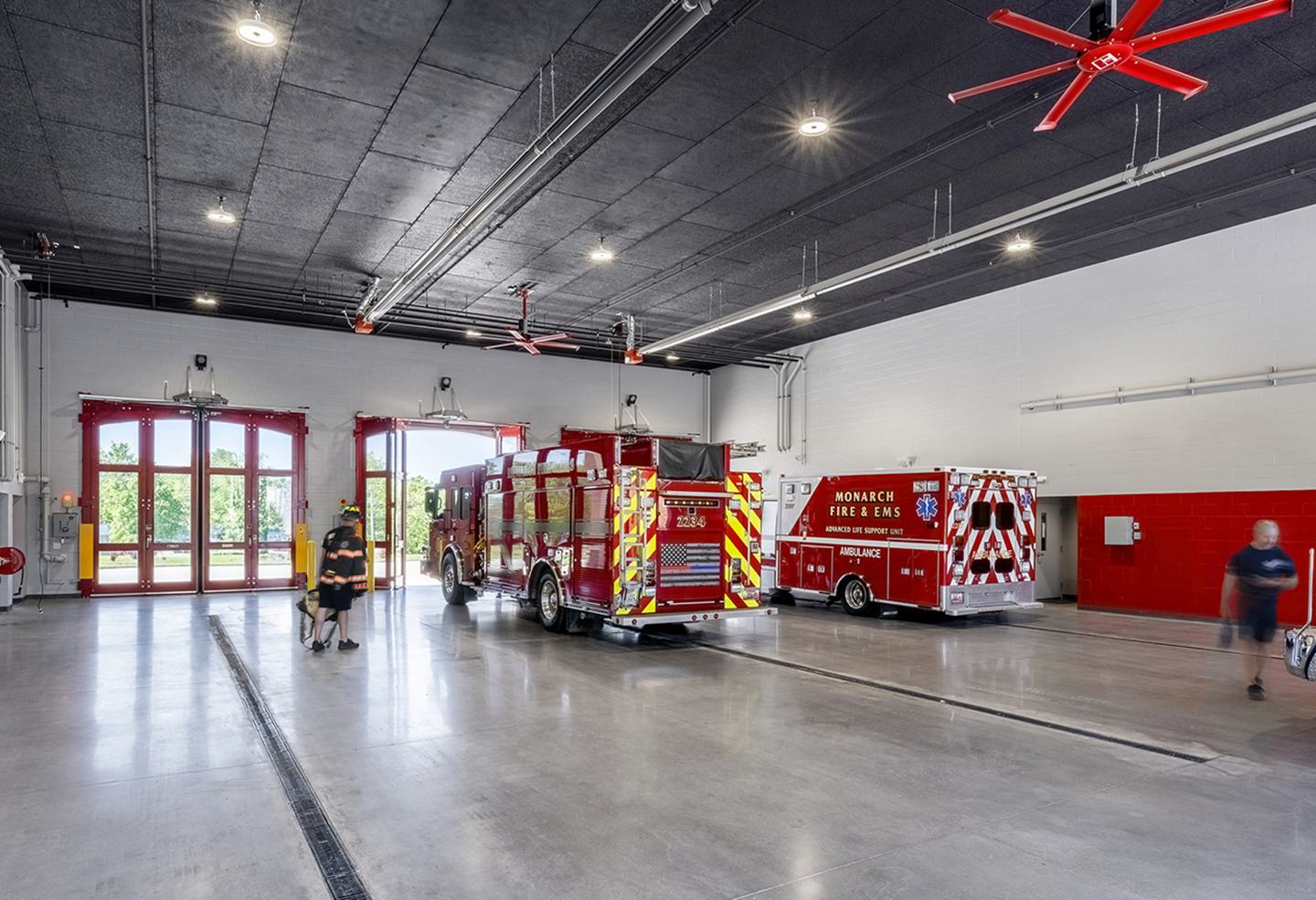 A spacious fire station interior with red and white fire trucks, large doors, and a clean, modern design. Ceiling fans enhance airflow.